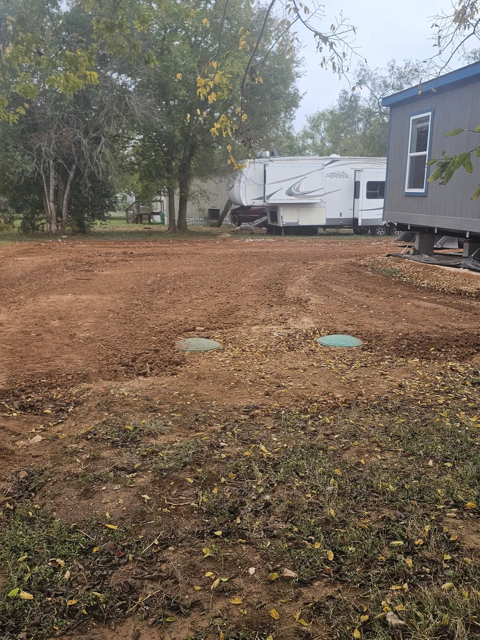 RV park site with graded dirt lot, two septic access covers, and parked RVs under trees